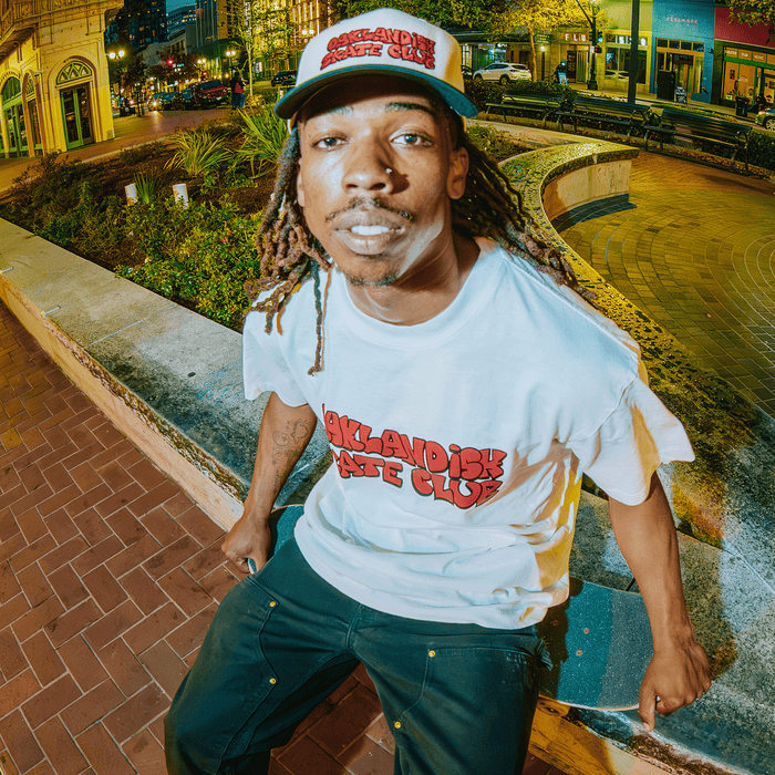 A man with long dreadlocks, a mustache, and beard sits on a curved concrete bench at night in the city, wearing an Oaklandish Skate Club 2-Tone Hat by Oaklandish and a white t-shirt, capturing skateboard culture under urban lights.