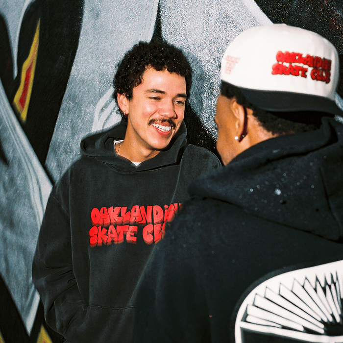 Two young men stand before a graffiti mural, smiling and chatting. Both wear black hoodies with "Oaklandish Skate Club" in red, while one rocks the Oaklandish Skate Club 2-Tone Hat, repping true skateboard culture.