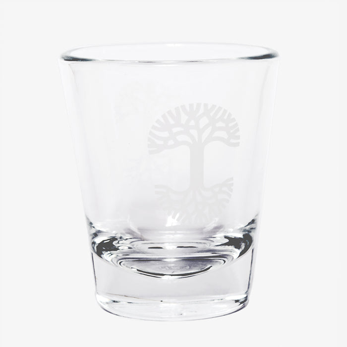 The Oaklandish Trees Shot Glass by Oaklandish is a clear glass tumbler with a thick, solid base, showcasing an elegant logo etching of a stylized tree with sprawling branches and roots, symbolizing the tree of life. This exquisite shot glass is empty and set against a plain white background.