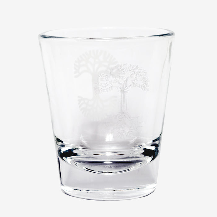 An Oaklandish Trees Shot Glass by Oaklandish is displayed against a plain white background. The clear glass features an etched tree design with intricate, branching roots and branches, forming a symmetrical and detailed pattern. The shot glass possesses a slight shine and reflection, suggesting it is clean and smooth.