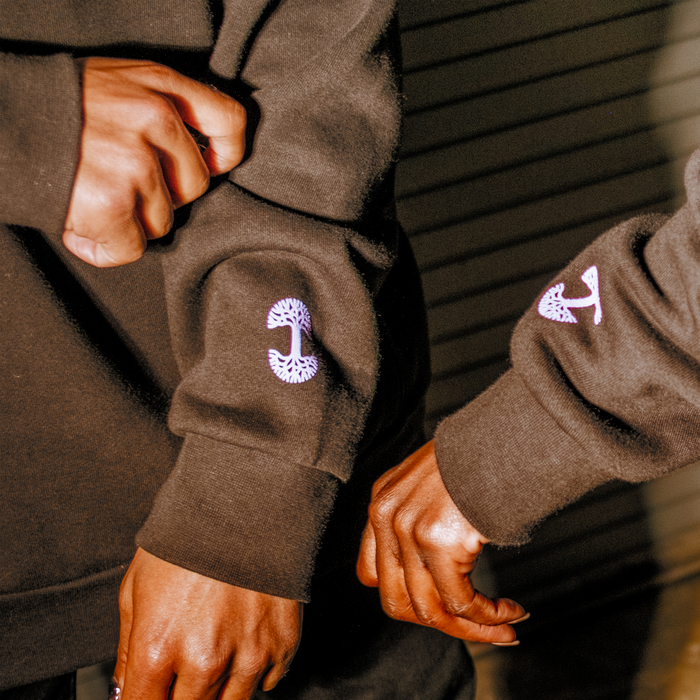 Two people wear GSV Odin Hoodies—unisex fleece sweatshirts with white and purple mushroom sleeve designs. One person grips a sleeve as their hands nearly meet, set against a blurred background with horizontal dark lines.