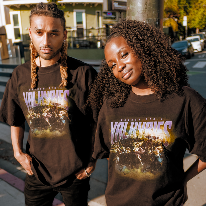 Two people stand outdoors in matching black GSV Odin Tees. The person on the left has long blond braids and a beard; the right, with curly hair, smiles as sunlight highlights their 100% cotton shirts against a city backdrop.