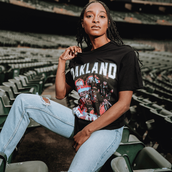 A woman with braided hair sits on green stadium seats, confidently showing off her black OFA Tour Tee by Oakland Roots SC, paired with ripped jeans as she faces the camera and proudly displays her love for the club.