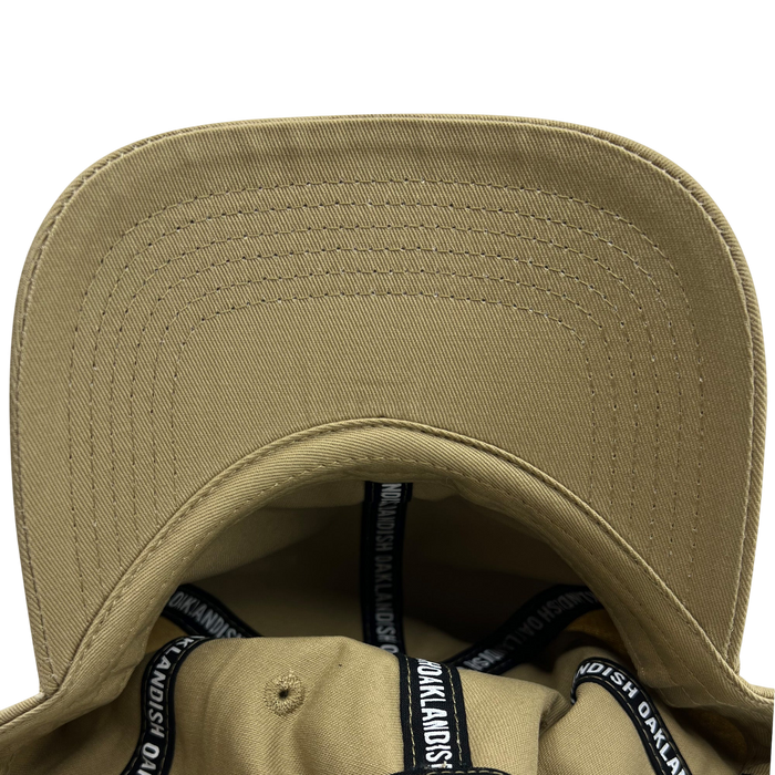 A close-up view of the inside of the Official Micro Logo Dad Hat by Oaklandish, showcasing its beige color, curved brim, and precise stitching. The inner lining is adorned with black bands featuring white text that repeatedly spells "Oaklandish" around the edges. The sturdy fabric and clear stitching are evident throughout, along with an adjustable strap for a perfect fit.