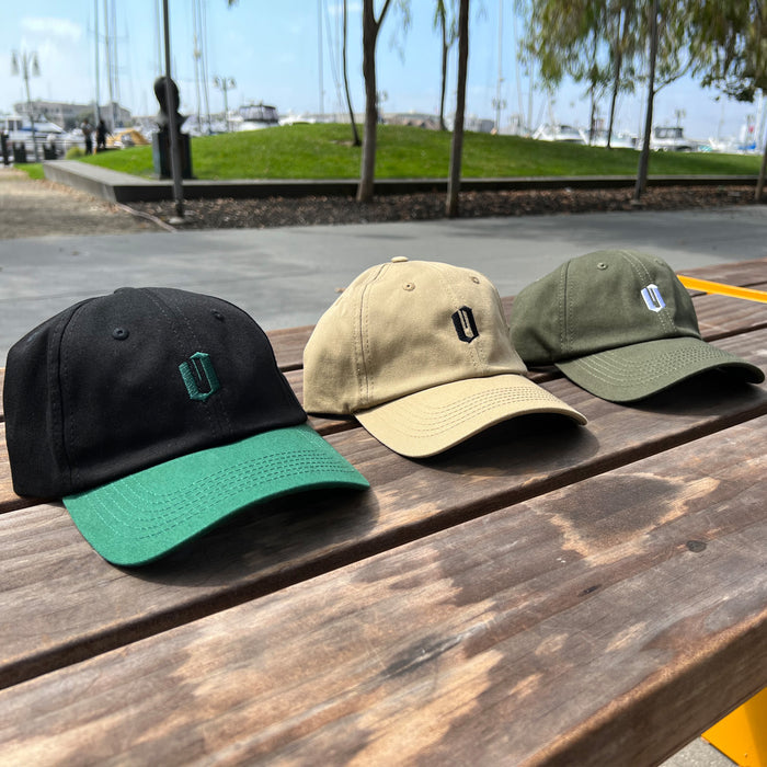 Three Oaklandish Official Micro Logo Dad Hats are displayed on a wooden surface. From left to right, the caps are black with a green brim, beige, and olive green. Each hat features a simple embroidered logo on the front. In the background, there is a marina with boats and trees visible. The adjustable straps add convenience to their stylish design.