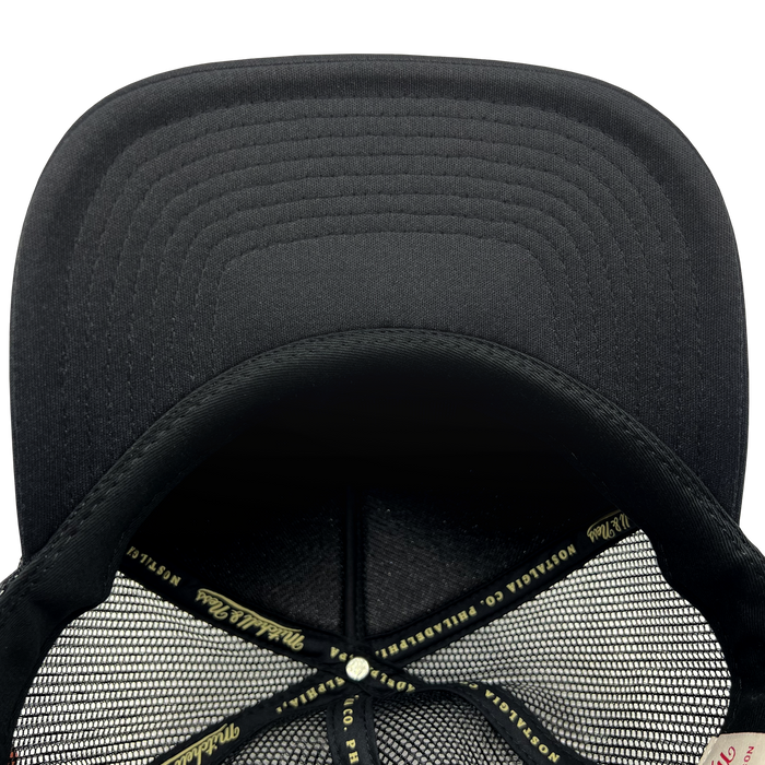 Close-up view of the interior of a black Official O Trucker baseball cap by Oaklandish. The inside features a mesh lining with crosshatched stitching and a central button. The cap has fabric labeled "PIPILA" and other branding on the inner band that runs around the base. The brim shows reinforced stitching.