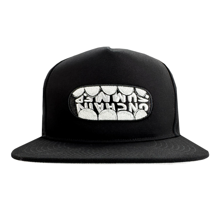 The OG Snapback Hat by Supported Brands is a black adjustable snapback featuring a front embroidered patch where stylized white teeth spell “UNCHAIN” across the upper row. Photo shows the hat on a plain white background.