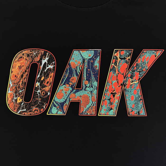 The Oil and Water Tee by Oaklandish features the word “OAK” in bold letters with vibrant marbled patterns on a black background, making it a striking classic-fit men’s t-shirt crafted from heavy-weight cotton.