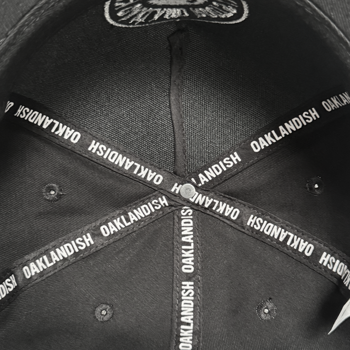 Inside view of the ORSC 5 Panel Hat by Oakland Roots SC, a top Oakland sports item. Intersecting fabric strips labeled “OAKLANDISH” form a star around a central button, with visible stitching and metal eyelets on black fabric.