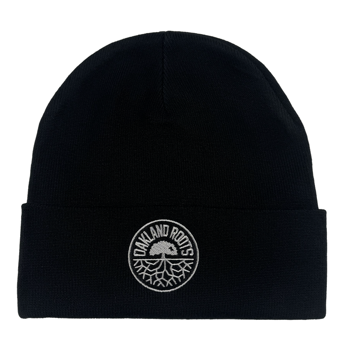 The ORSC Beanie by Oakland Roots SC is a black knit hat with a folded brim and a circular white patch on the front featuring "OAKLAND ROOTS" and the club’s tree logo. The background is plain white.