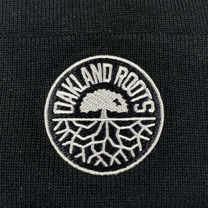 A close-up of the black ORSC Beanie by Oakland Roots SC, showcasing a circular white-embroidered patch with “OAKLAND ROOTS” and a solid-canopy tree with exposed roots.