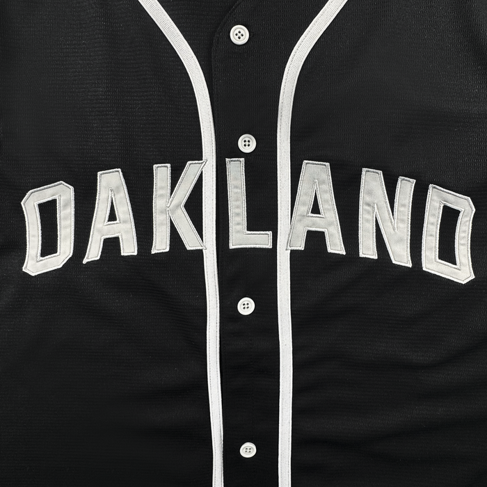 A close-up of the ORSC Button Up Jersey by Oakland Roots SC, featuring bold white "OAKLAND" lettering outlined in silver across the chest. This black jersey showcases the signature Oaklandish Lifestyle style with white buttons down the front and white trim along the button panel.