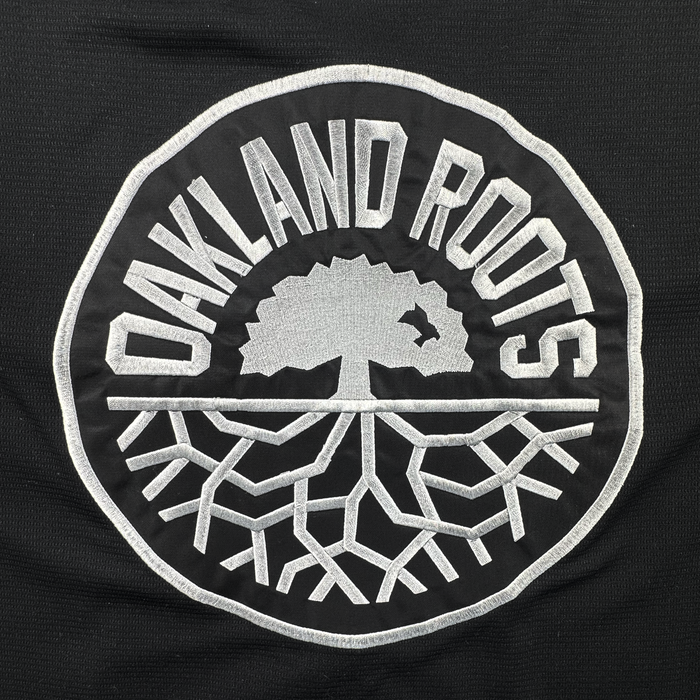 Circular black patch with "OAKLAND ROOTS" in bold white letters. At the center is a white tree with spreading roots and a small bird flying near the branches. Inspired by the Oakland Roots SC, this bold, graphic emblem is perfect for showing local pride on the ORSC Button Up Jersey by Oakland Roots SC.