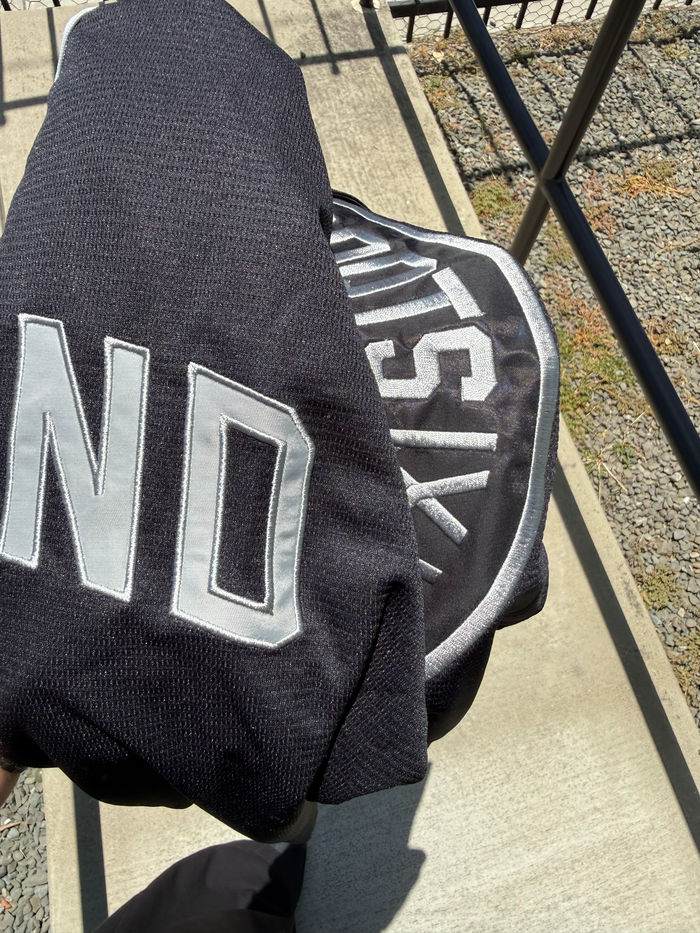 A person holds a folded ORSC Button Up Jersey by Oakland Roots SC, featuring bold white lettering and a partial circular logo, while standing on a sunlit concrete path beside a metal rail and gravel.