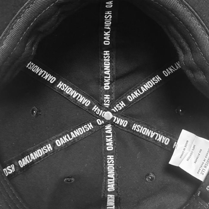 Black and white photo of the inside of an ORSC Dad Hat by Oakland Roots SC. Inner bands display "OAKLANDISH" in white text radiating from the center button, with a white clothing tag attached on the right side.