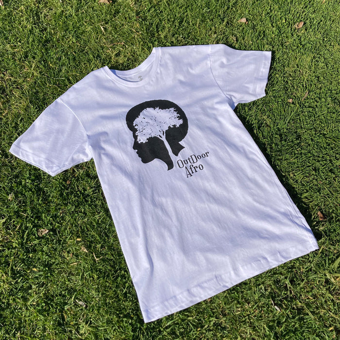 The Outdoor Afro Signature Tee by Partner lies on a bed of grass, showcasing a black silhouette of a person's head with a tree forming the hair. "Outdoor Afro" is printed in black beneath the tree. The design stands out against the vibrant green grass, highlighting an outdoors theme.