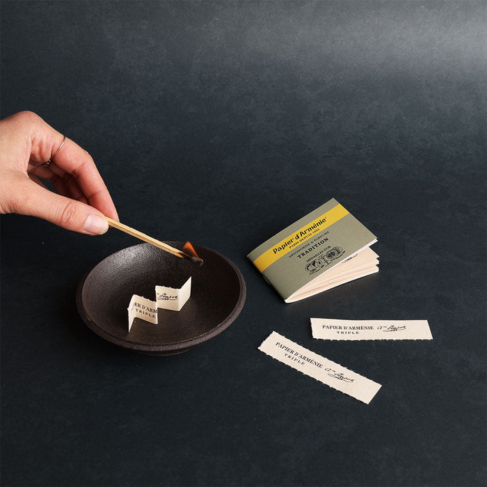 Hand lighting a match with a small book of incense papers on a dark surface