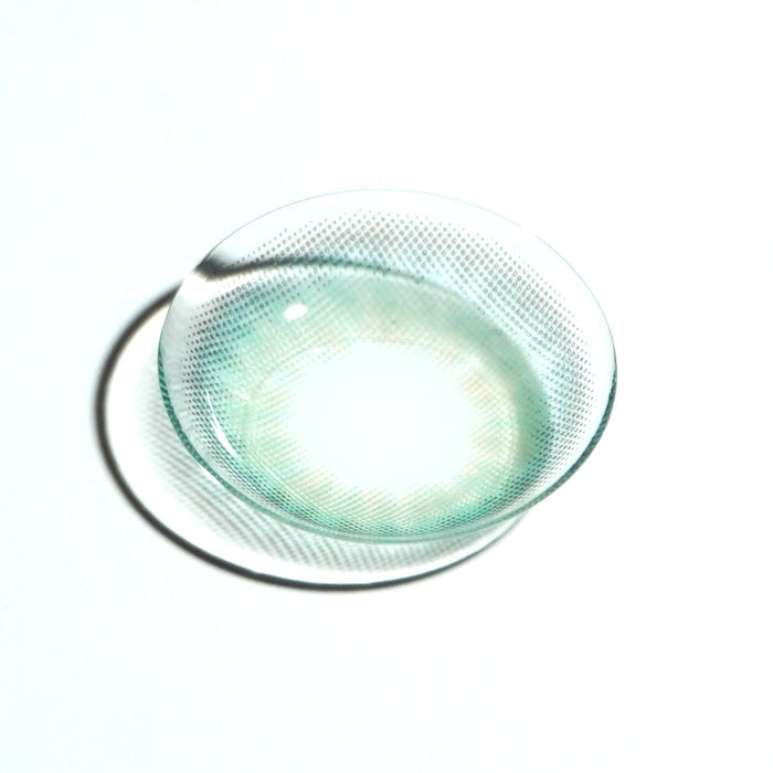 A single EyeCandys Pink Label Mallorca Blue contact lens, featuring a translucent blue with subtle green tint and dotted pattern, rests on a white surface and casts a soft shadow.
