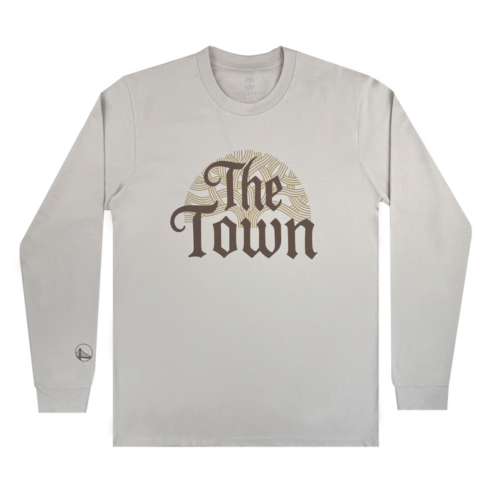 A light gray long-sleeve Pirate Black-Letter LS Tee by Other, featuring "The Town" in bold, ornate brown letters across the chest over a circular gold sunburst design. A small logo appears near the left sleeve cuff. The shirt is laid flat on a plain white background.
