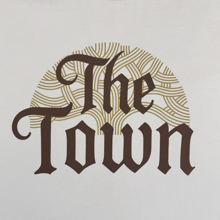 Stylized text reading "The Town" appears in bold, dark letters on a plain white background. Behind the text, a semicircle pattern of tan curved lines suggests tree rings or abstract topography—a signature design featured on the Pirate Black-Letter LS Tee by Other and evocative of the Warriors x Oaklandish collaboration from the Town Collection.