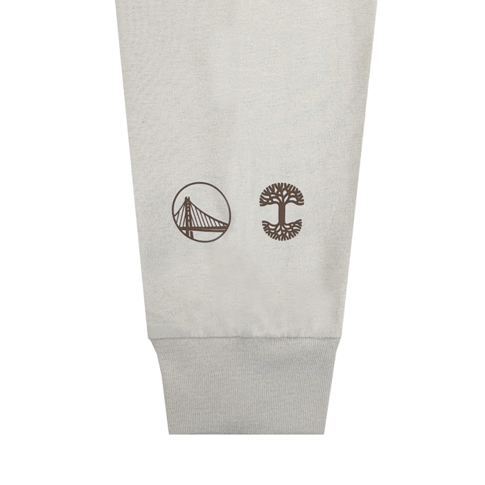 A close-up of a light gray fabric cuff from the Pirate Black-Letter LS Tee by Other shows two brown logos: a stylized suspension bridge in a circle on the left, and a tree with wide branches and roots, also in a circle, on the right. The background is plain white.