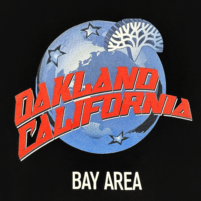 The Planet Oakland Tee by Oaklandish features a blue Earth with white stars and a stylized tree. Bold red “OAKLAND CALIFORNIA” spans the globe, with “BAY AREA” in white below, all on soft 100% cotton.