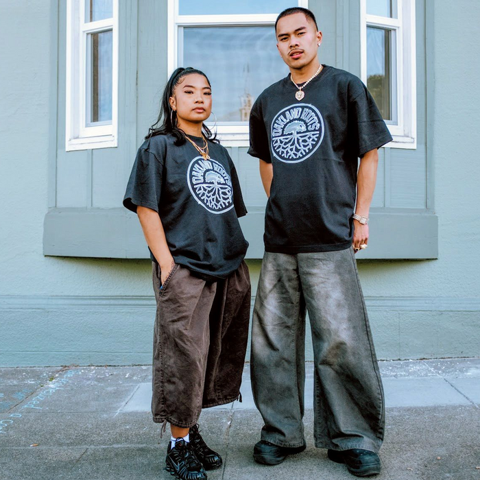 Two people stand outside a green wall with a window, each wearing Oakland Roots SC Platinum Crest Tees and loose, wide-legged pants. Looking confident with hands in pockets, their relaxed outfits exude effortless style.