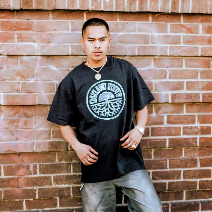 A man with short hair and a mustache stands against a brick wall wearing the Oakland Roots SC Platinum Crest Tee, gold jewelry, and gray pants. His left hand rests on his pocket as he looks directly at the camera with a serious expression.