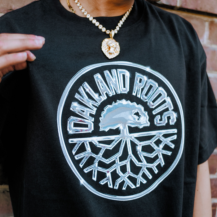 Wearing the Oakland Roots SC Platinum Crest Tee, featuring a bold "OAKLAND ROOTS" logo with a tree and roots, plus a gold lion head chain. The brick wall background and partial frame highlight Pride for Roots SC.