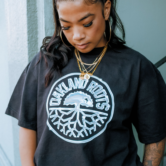 A woman with hoop earrings and layered gold necklaces looks down, partially revealing her tattooed arm. She wears the Oakland Roots SC Platinum Crest Tee, featuring a large white tree logo with roots inside a circle.
