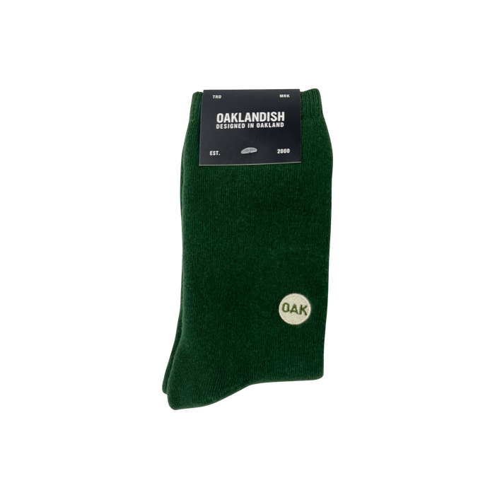 A pair of dark green Plush Oak Socks by Oaklandish are neatly folded and secured with a black label at the top. The label reads “OAKLANDISH,” “Designed in Oakland,” “Est. 2000.” These socks feature a small white circle emblem on the side, containing the letters “OAK” in green.