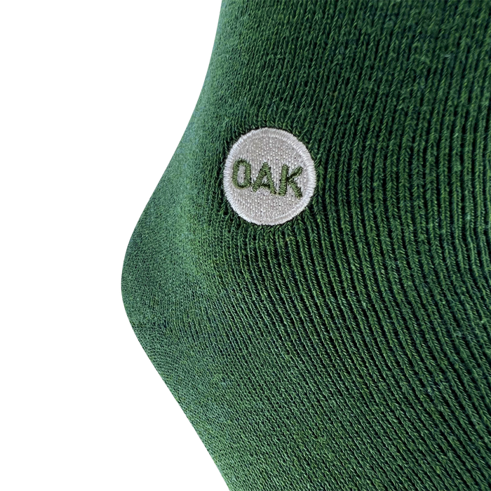 A close-up of a green fabric, likely from an Oaklandish Plush Oak Sock, with a textured, knitted pattern. There is a small white circular patch on the fabric with the uppercase letters "OAK" embroidered in green inside the patch. The background is plain white—plush socks for happy feet.