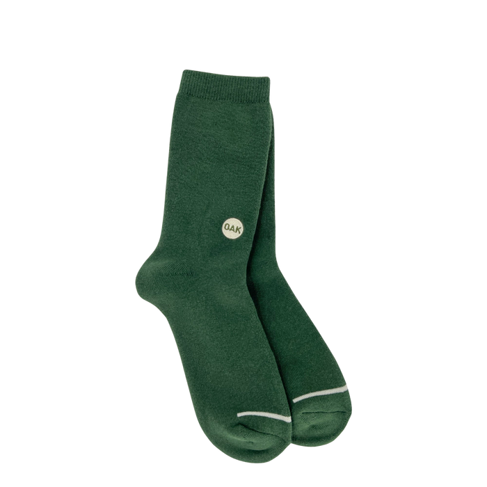 A pair of Oaklandish Plush Oak Socks in dark green is showcased against a plain white background. Each sock is adorned with a small circular Oak logo marked with the letters "QAK" near the top outer side. These plush socks feature a subtle white outline at the toe area and have a ribbed cuff at the top, ensuring happy feet all day long.