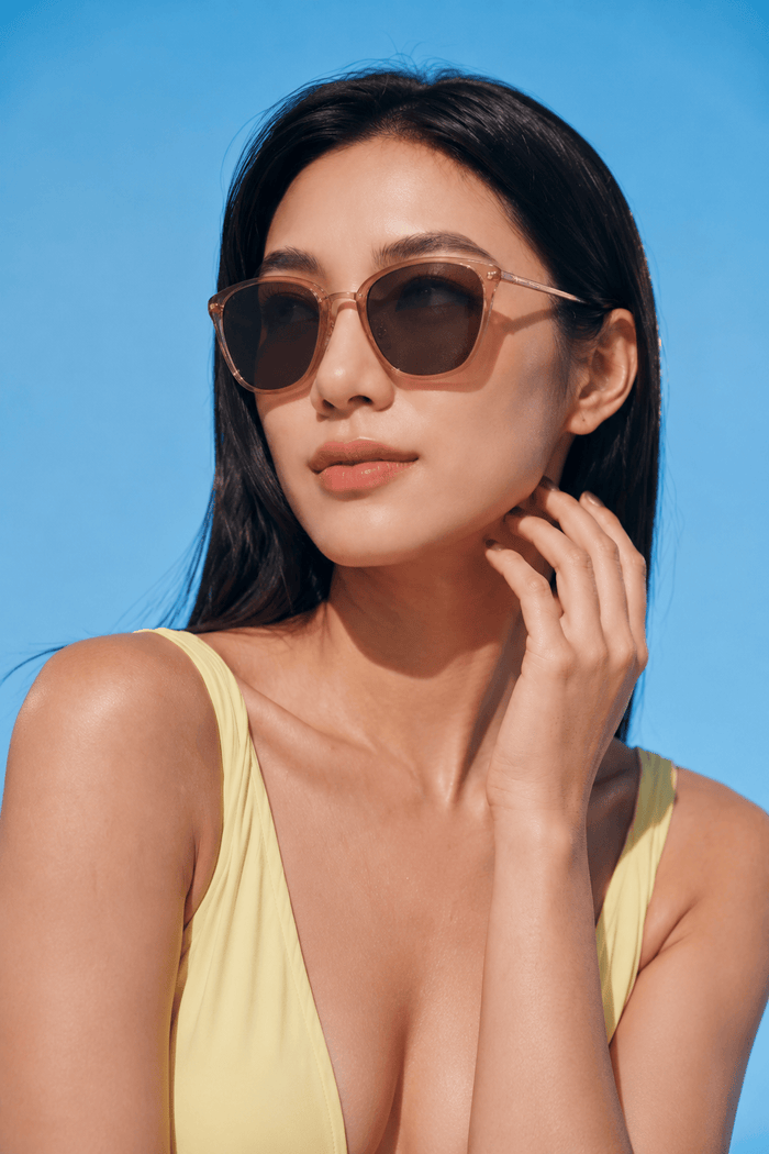 A woman in a yellow swimsuit poses against a bright blue sky, wearing EyeCandys Posh Square full-frame glasses, touching her face and gazing into the distance with a relaxed expression.