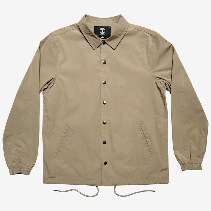 The Oaklandish Premium Coaches Jacket is a beige, long-sleeve jacket featuring a collar and elastic cuffs. This water-resistant jacket includes a drawstring hem, diagonal front pockets, and black snap buttons down the front. The interior label displays a white anchor symbol on a black background. The men's sizing jacket is shown laid flat against a white backdrop.