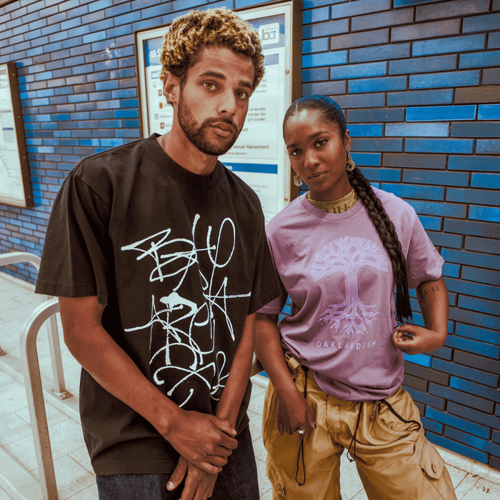 A man in a men's black heavy-weight cotton tee and a woman in a lavender Oaklandish Presto Tee with braided hair and tan cargo pants stand together by blue-tiled walls and transit maps.