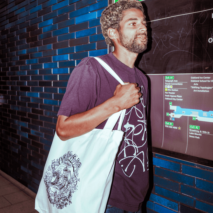 A man with curly hair and a beard, wearing an Oaklandish Presto Tee—men's black t-shirt with white abstract designs in a classic fit—stands near a green and black transit map, carrying a heavy-weight cotton tote bag with detailed black artwork.