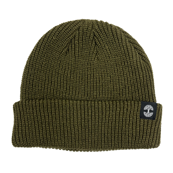 The Ramos Beanie by Oaklandish is a green knit short beanie with a folded brim. It features a black patch displaying a white tree logo and is made from woven acrylic. The ribbed texture adds to its cozy look, and the headwear piece is photographed laying flat on a white background.