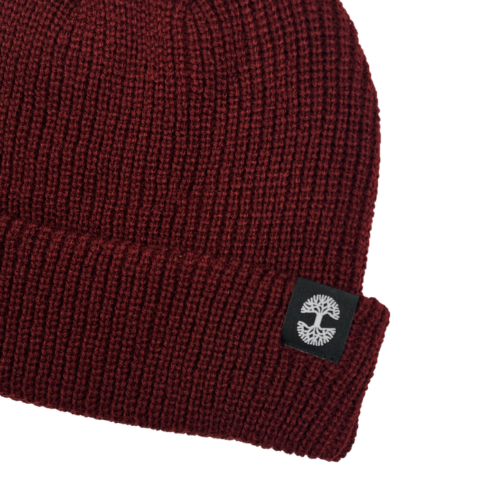 A close-up of the Ramos Beanie by Oaklandish, a dark red knit crafted from woven acrylic with a folded brim. The brim showcases a black patch featuring a white abstract tree logo on the side. This stylish piece of headwear has a textured, ribbed pattern and is set against a plain, light gray background.