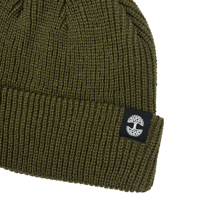 The Ramos Beanie by Oaklandish is an olive green knit hat made from woven acrylic. It showcases a folded brim adorned with a black fabric patch that features a white tree emblem. The beanie has a ribbed texture, contributing to its cozy and warm look.