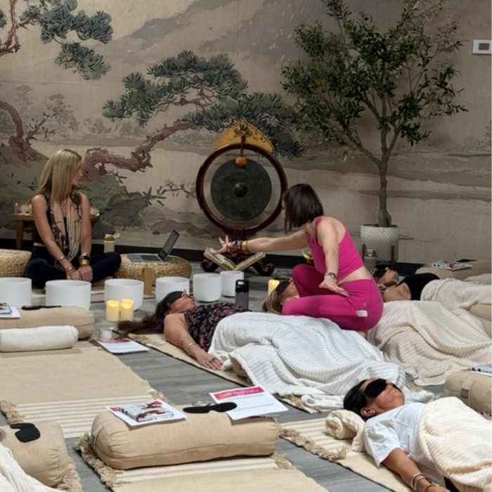 Participants relax on cushioned mats during a restorative sound bath as a facilitator plays a large gong in a serene mural filled wellness space by BuDhaGirl