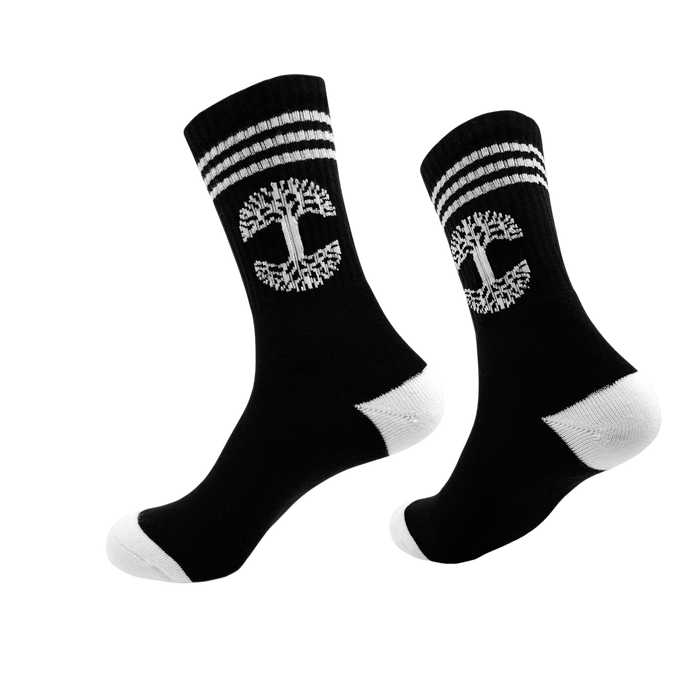 A pair of Retro Classic Socks from Oaklandish, exceptionally comfy in black, featuring a white tree emblem and two horizontal white stripes near the top. The socks have unique white heels and toes, displayed with one sock slightly in front of the other against a plain white background.