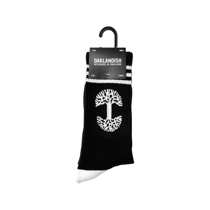 A pair of exceptionally comfy Retro Classic Socks by Oaklandish, featuring a black base with white accents. These socks showcase a white tree symbol in the center, white toes, and two white stripes near the top. The packaging label proudly displays "Oaklandish," along with "Designed in Oakland" and "EST. 2000.