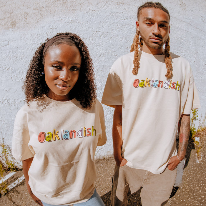 A man and woman stand before a textured white wall, both wearing Oaklandish Scrawl Tees—classic fit, light-colored t-shirts featuring “Oaklandish” in colorful outlined letters. The men's cotton tee stands out for its comfortable style.