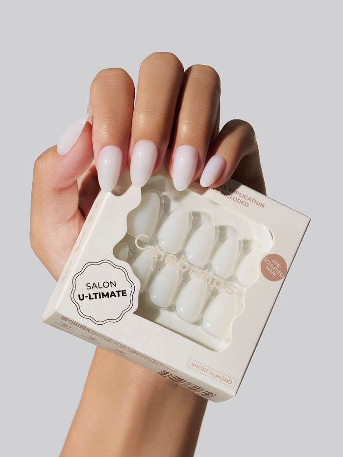 Hand wearing White Salon U-ltimate short almond press-on nails, holding a box of the nails on a white background.
