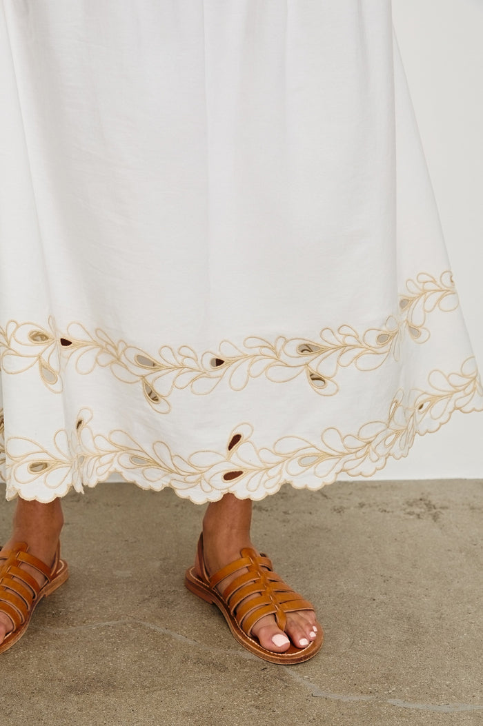 Detail image of model wearing a white, cotton poplin midi skirt with a lightly gathered waist that cinches and drops at the hips. Details include beige embroidered eyelets at the hem. Worn with brown sandals.
