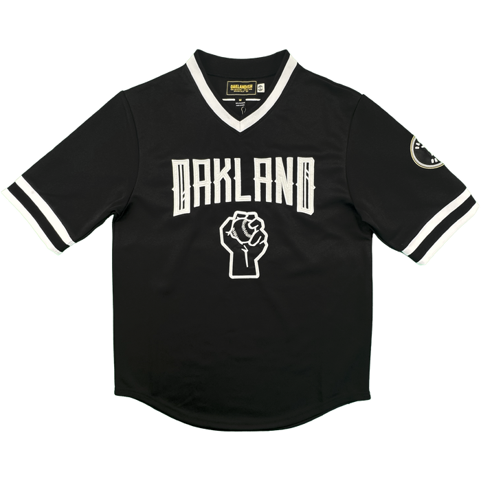 The Stands Up Jersey by Oakland Ballers is a black short-sleeve jersey with white-trimmed collar and sleeves, "OAKLAND" in bold white above a raised fist graphic, white sleeve stripes, and a circular patch on the left sleeve.