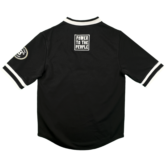 The Stands Up Jersey by Oakland Ballers is a black short-sleeved shirt with white trim, a “POWER TO THE PEOPLE” patch on the upper back, and an Oakland Ballers circular patch on the left sleeve, shown on a plain white background.