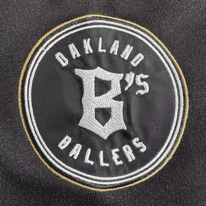 The Stands Up Jersey by Oakland Ballers features "Oakland Ballers" in white text encircling a large gold "B" and "'75" on a black background, bordered with white and gold rings.