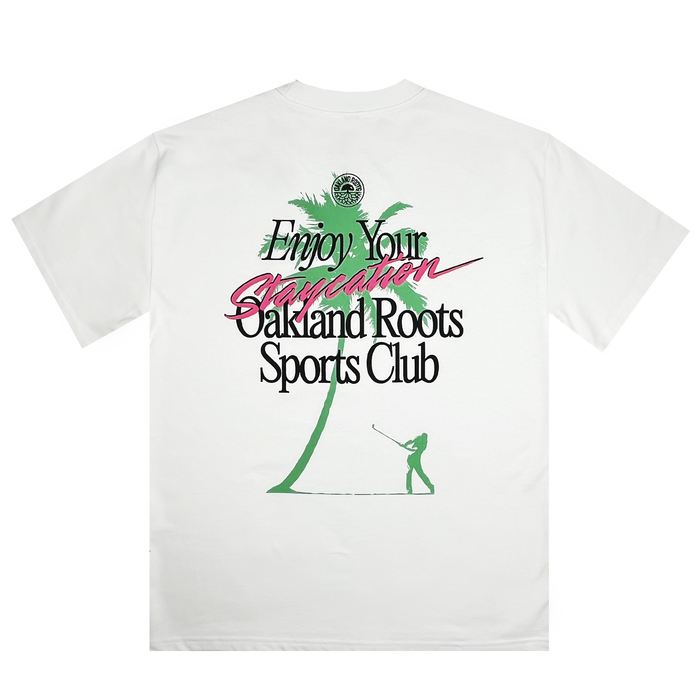 The Staycation Heavy Tee by Oakland Roots SC features a boxy fit and showcases a green palm tree alongside a silhouette of a person golfing. The design includes the text "Enjoy Your Staycation" in teal and pink colors, with "Oakland Roots Sports Club" written below in black. As part of the Oaklandish collection, this shirt also supports social good initiatives.